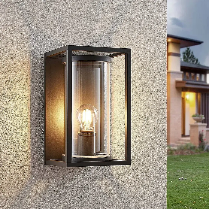 Lucande Außenwandleuchte Brienne - alu / grau / zink, IP54 - Schwarze Außenwandleuchte mit klarem Glaszylinder, beleuchtet eine strukturierte Wand neben einem Haus.