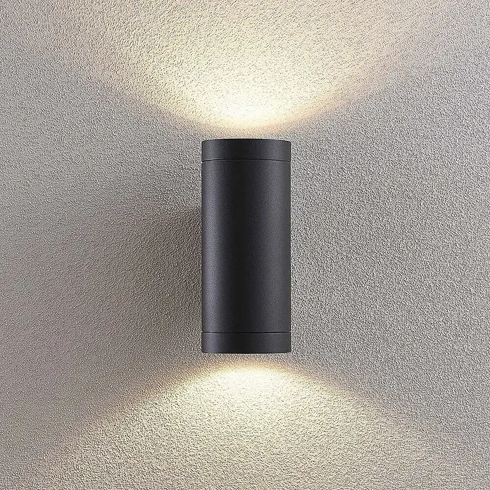 Lucande Außenwandleuchte Thomke - alu / grau / zink, alu / grau / zink, IP65 - Schwarze zylindrische Up-Down-Wandleuchte an einer strukturierten Wand, die warmes Licht nach oben und unten wirft.