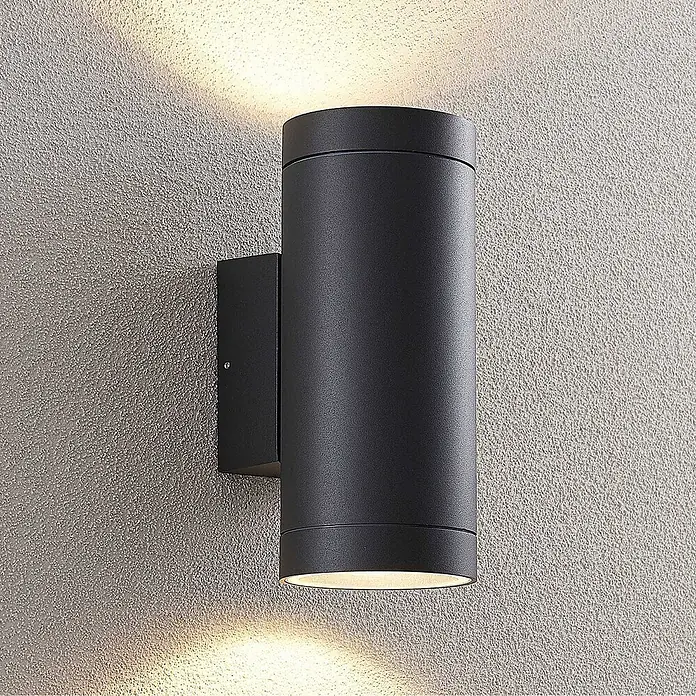 Lucande Außenwandleuchte Thomke - alu / grau / zink, alu / grau / zink, IP65 - Schwarze zylindrische Up-Down-Wandleuchte, die warmes Licht auf eine strukturierte Wand wirft.