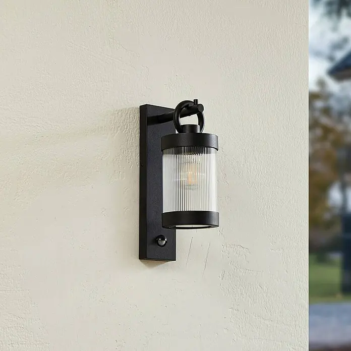 Lindby Außenwandleuchte Yrene - schwarz, schwarz, IP44 - Schwarze Außenwandleuchte mit geripptem Glasschirm und Bewegungssensor an strukturierter Wand.