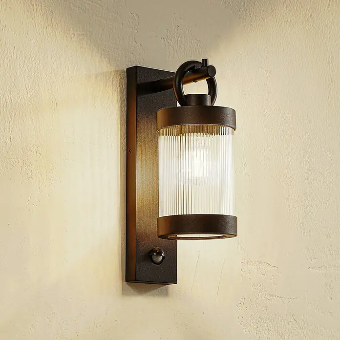 Lindby Außenwandleuchte Yrene - schwarz, schwarz, IP44 - Dunkelbronze Außenwandleuchte mit geriffeltem Glas und Bewegungssensor an einer texturierten Wand.
