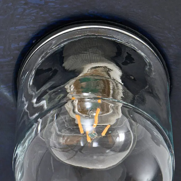 Lindby Außenwandleuchte Eloretta - schwarz, schwarz, IP44 - Eine klare Glaslaterne mit einer LED-Glühbirne mit leuchtenden orangefarbenen Filamenten, montiert an einer dunkelblauen strukturierten Wand.