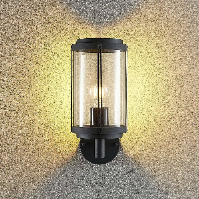 Lindby Außenwandleuchte Ojali - schwarz, IP44 - Schwarze zylindrische Außenwandleuchte mit Klarglas und Filamentlampe, beleuchtet eine strukturierte Wand.