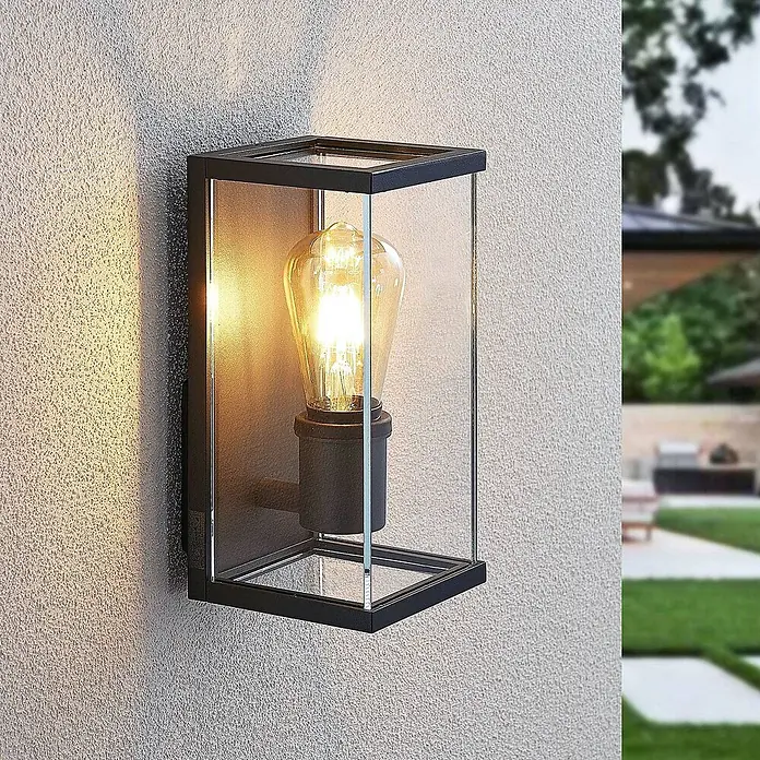 Lindby Außenwandleuchte Susanne - 13 x 12 x 25 cm, schwarz, IP44 - Schwarze Metall-Glas-Außenwandleuchte mit Vintage-Glühbirne, montiert an einer strukturierten Wand im Garten.