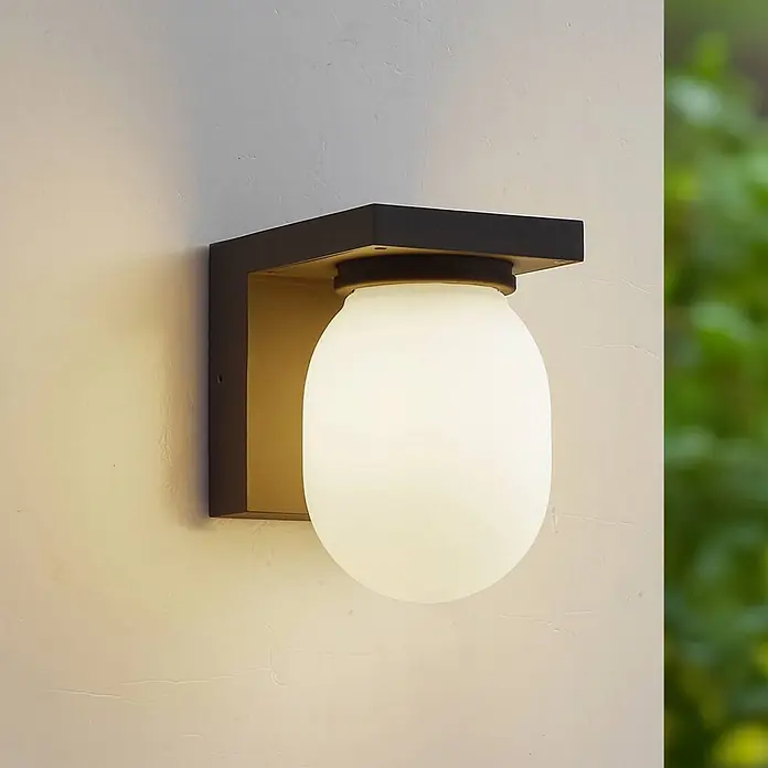 Lucande Außenwandleuchte Ovaali - schwarz, IP65 - Moderne Außenwandleuchte mit dunkler rechteckiger Halterung und mattweißer Glaskugel, an einer texturierten Wand befestigt.