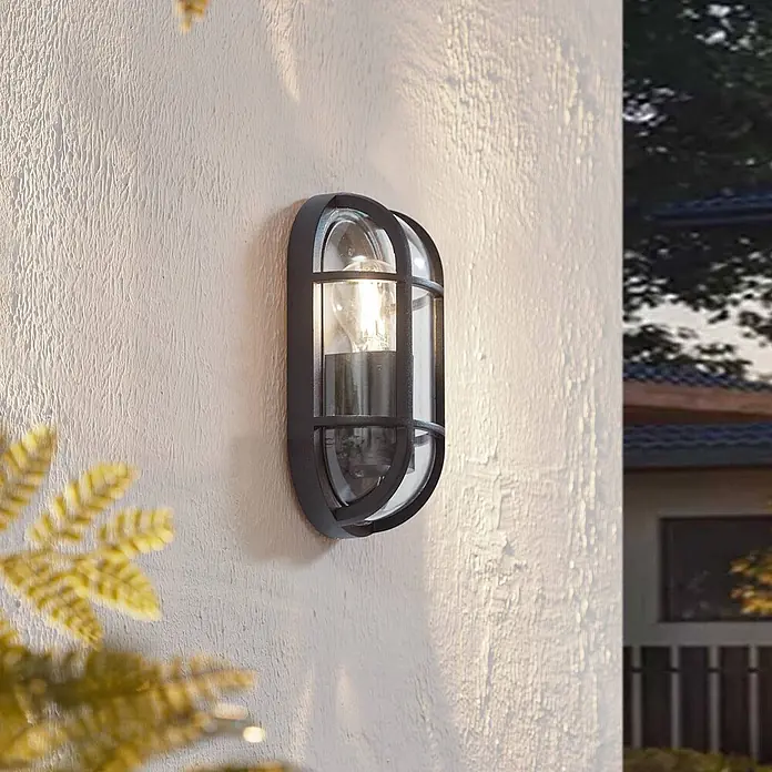 Lucande Außenwandleuchte Serine - schwarz, klar / transparent, IP65 - Schwarze ovale Außenwandleuchte mit Gitterdesign und Klarglas, montiert an einer strukturierten Wand und beleuchtet.