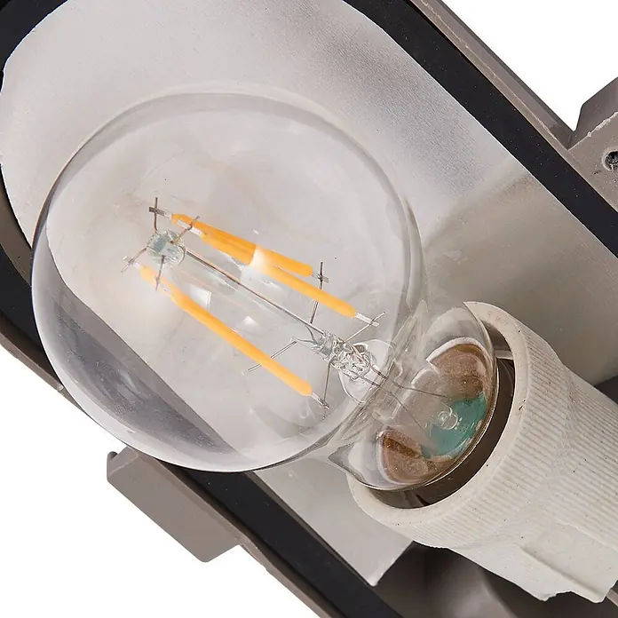 Prios Außenwandleuchte Kirgo - 11,3 x 16 x 12,5 cm, alu / grau / zink, IP44 - Nahaufnahme einer klaren LED-Filamentlampe mit orangefarbenen Filamenten, in einer weißen Keramikfassung einer silbergrauen Metallleuchte.