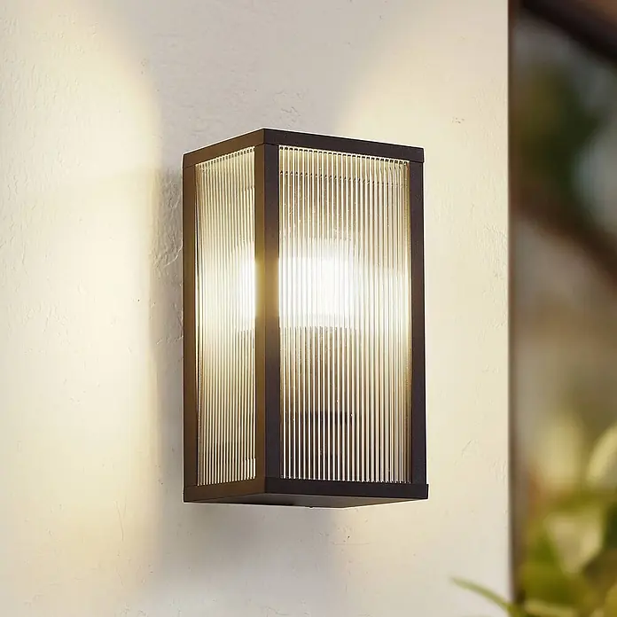 Lindby Außenwandleuchte Peldar - 13,5 x 14 x 26 cm, schwarz, IP44 - Beleuchtete Outdoor-Wandleuchte aus dunklem Metall mit gerippten Glasscheiben, an einer strukturierten Wand.
