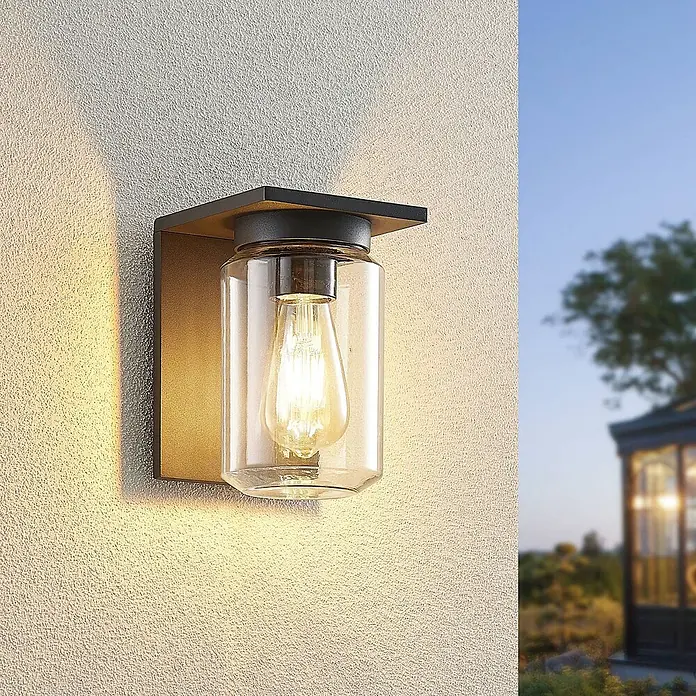 Lucande Außenwandleuchte Semka - schwarz, klar / transparent, IP54 - Außenwandleuchte mit dunklem Metallrahmen, Messingakzent und klarem Glaszylinder um eine Edison-Glühbirne, an einer strukturierten Wand montiert.