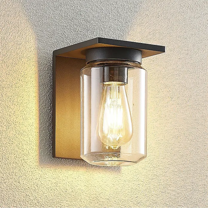 Lucande Außenwandleuchte Semka - schwarz, klar / transparent, IP54 - Wandleuchte aus schwarzem Metall und Klarglas mit Edison-Birne, die eine strukturierte Wand beleuchtet.