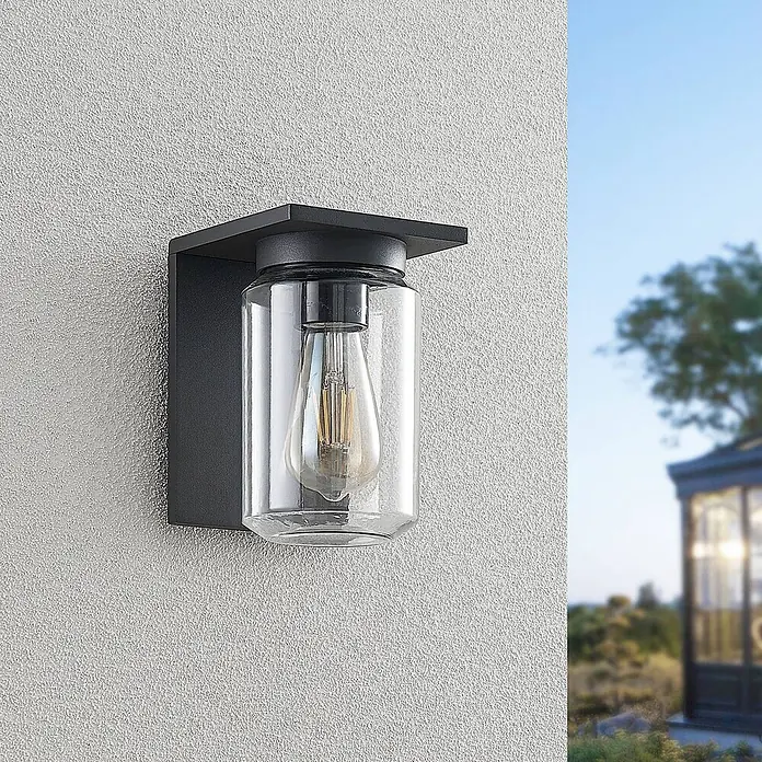 Lucande Außenwandleuchte Semka - schwarz, klar / transparent, IP54 - Schwarze Außenwandleuchte mit klarem Glaszylinder und sichtbarer Glühbirne, an einer strukturierten Wand.