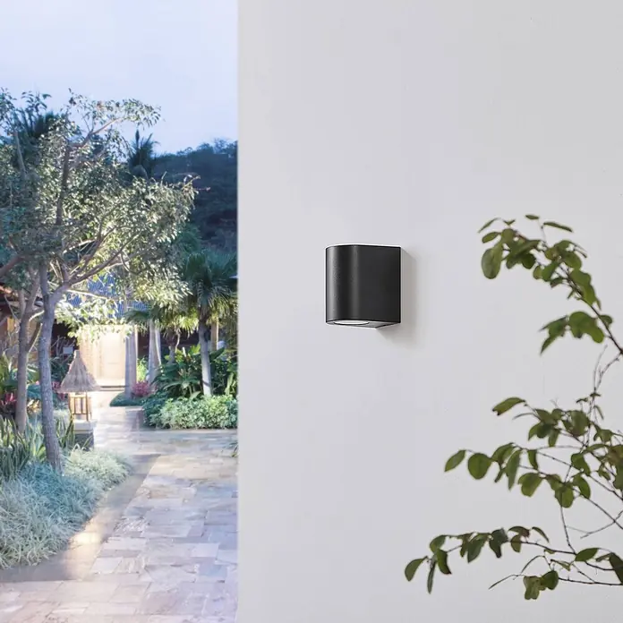 Prios Außenwandleuchte Irfan - 8,9 x 6,8 x 10 cm, schwarz, IP44 - Schwarze rechteckige Außenwandleuchte, beleuchtet einen Garten.