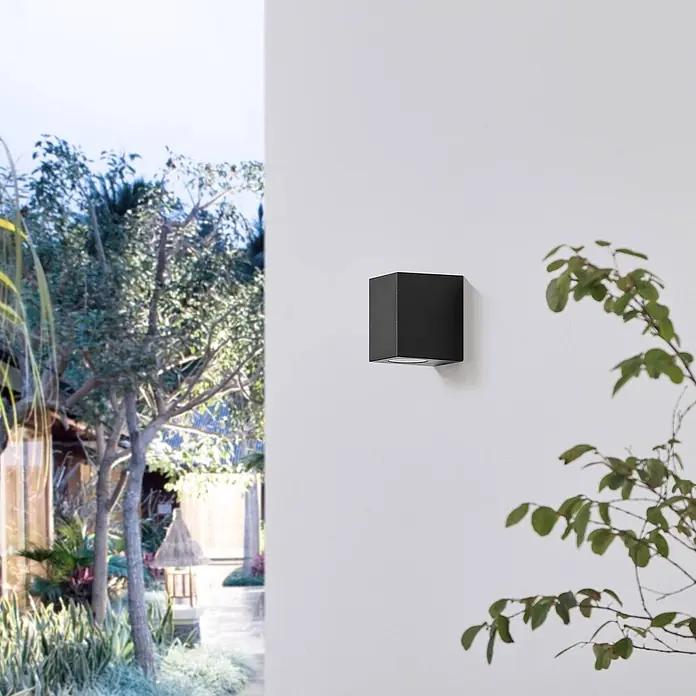 Prios Außenwandleuchte Irfan - 8,9 x 6,8 x 10 cm, schwarz, IP44 - Schwarze würfelförmige Wandleuchte an einer weißen Wand, die nach unten in einem Außenbereich beleuchtet.