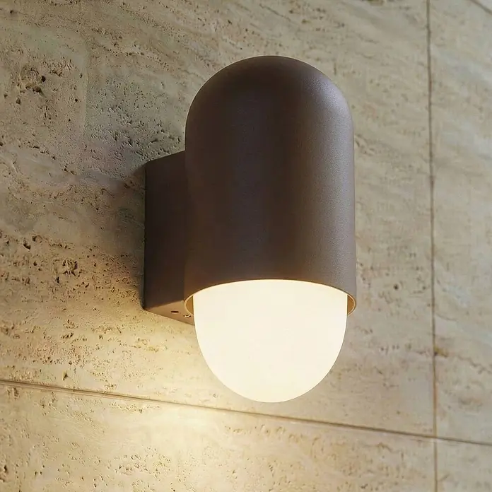 Lindby Außenwandleuchte Pilva - 13,3 x 10,5 x 21,5 cm, braun / rost, weiß / opal, IP44 - Dunkelbraune Außenwandleuchte mit weißem, leuchtendem Diffusor, an einer strukturierten Steinwand montiert.