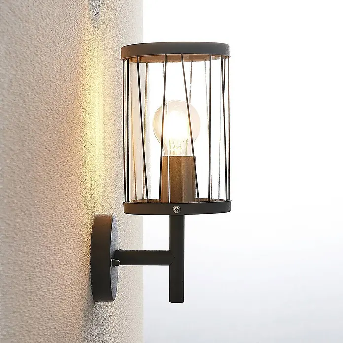 Lindby Außenwandleuchte Yonan - schwarz, schwarz, IP44 - Schwarze Wandleuchte aus Metall mit zylindrischem Klarglasschirm und sichtbarer Glühbirne, an strukturierter Wand montiert.