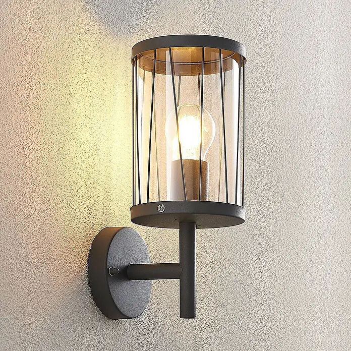 Lindby Außenwandleuchte Yonan - schwarz, schwarz, IP44 - Dunkelgraue Metall-Wandleuchte mit zylindrischem Glasschirm und sichtbarer Glühbirne, wirft warmes Licht auf eine strukturierte Wand.