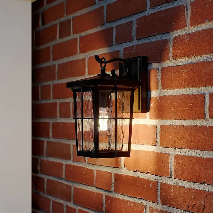 Lindby Außenwandleuchte Charmaine - 18,6 x 15,3 x 28,6 cm, braun / rost, braun / rost, IP23 - Schwarze Außenwandleuchte aus Metall mit strukturiertem Klarglas, beleuchtet eine Ziegelwand.
