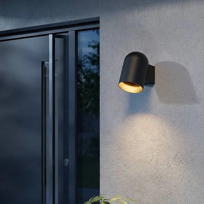 Lindby Wandleuchte aussen in schwarz, Aussenlampe Wand -  - Schwarze Wandleuchte aus Metall, nach unten gerichtet, erhellt eine strukturierte Wand neben einer dunklen Tür.