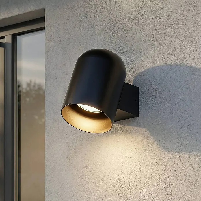 Lindby Wandleuchte aussen in schwarz, Aussenlampe Wand -  - Schwarze Außenwandleuchte mit goldenem Inneren, beleuchtet eine texturierte Wand neben einem Fenster.