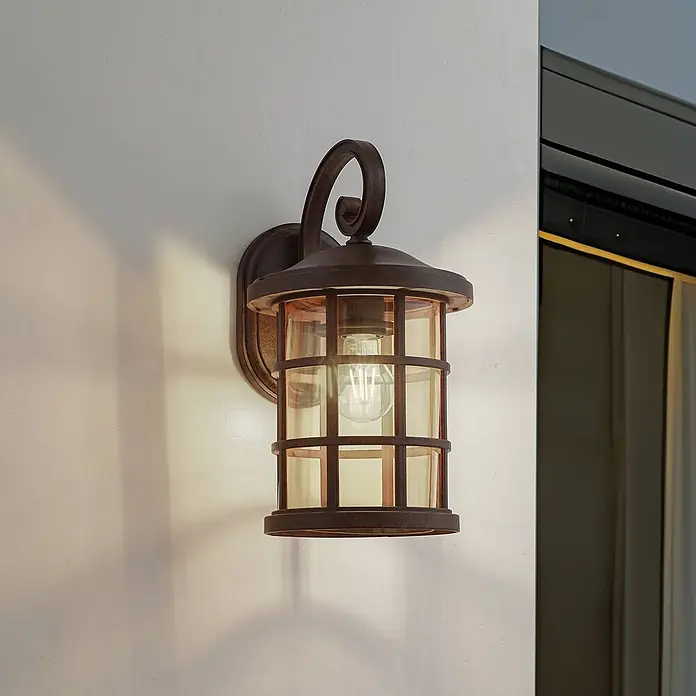 Lindby Wandleuchte aussen in rost, amber getönt, Aussenlampe Wand -  - Bronzefarbene Wandleuchte für den Außenbereich mit dekorativem Schnörkelarm und bernsteinfarbenem Glas im Käfigdesign, an einem Gebäude montiert.