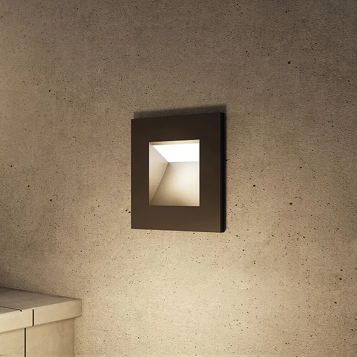 Arcchio Außenwandleuchte Zamo - schwarz, IP65 - Rechteckige, bronzefarbene Einbauwandleuchte an Betonwand, strahlt warmweißes Licht nach unten.