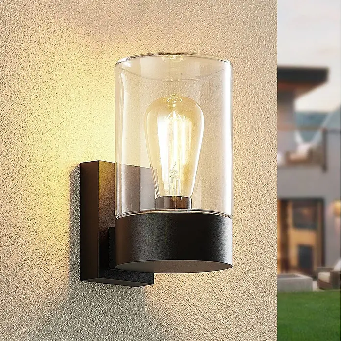Arcchio Außenwandleuchte Liljana - schwarz, klar / transparent, IP65 - Schwarze Außenwandleuchte mit klarem Glaszylinder und Edison-Glühbirne an einer strukturierten Wand.