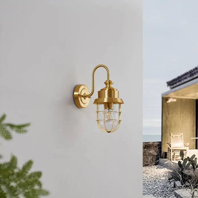 Lindby Außenwandleuchte Adalie - 21,5 x 13,7 x 31 cm, messing / gold, IP44 - Messingfarbene Outdoor-Wandleuchte mit einem Käfigglas-Schirm, an einer hellen Wand im Außenbereich.