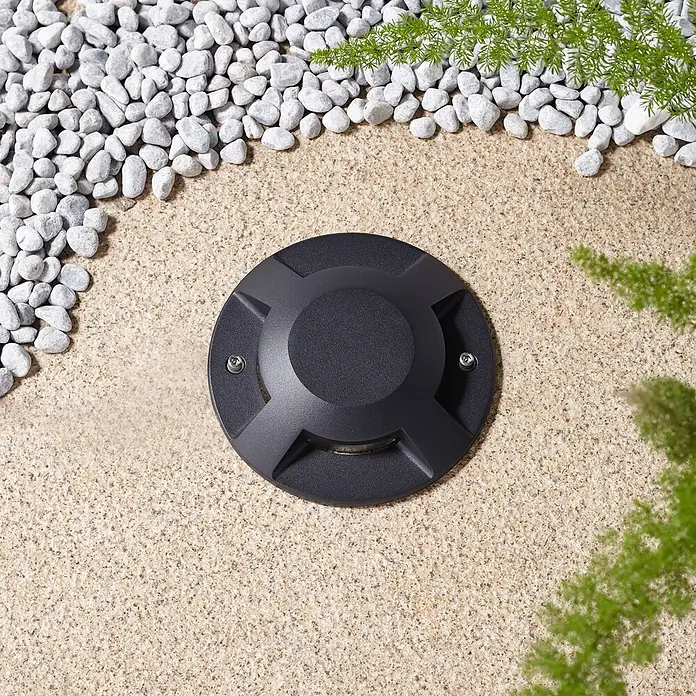 Lucande LED-Gartenstrahler Jeffrey - 3.000 K - Dunkelgraue kreisförmige Bodeneinbauleuchte in gesprenkelter beiger Oberfläche, umgeben von weißem Kies und grünem Laub.
