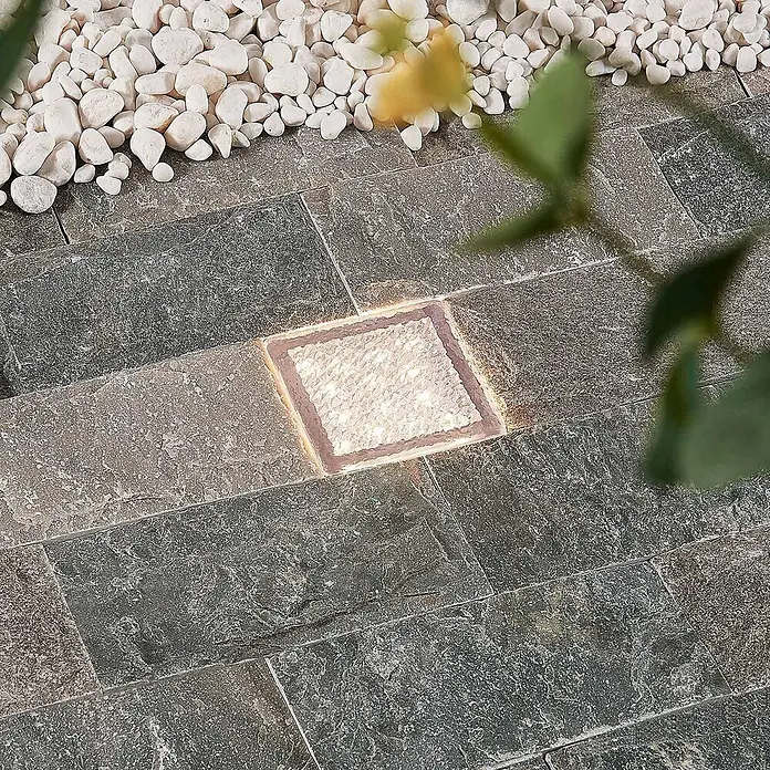 Prios LED-Gartenstrahler Ewgenie - 3.000 K, 10 x 10 x 6 cm - Quadratische, beleuchtete Außenbodenleuchte in dunkelgrauen Pflastersteinen, neben weißen Kieseln.