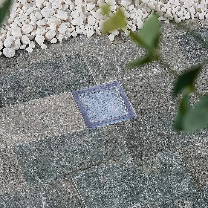 Prios LED-Gartenstrahler Ewgenie - 3.000 K, 10 x 10 x 6 cm - Quadratische Bodenleuchte, in einen gepflasterten Außenweg eingelassen, umgeben von weißen Zierkieseln und Grün.