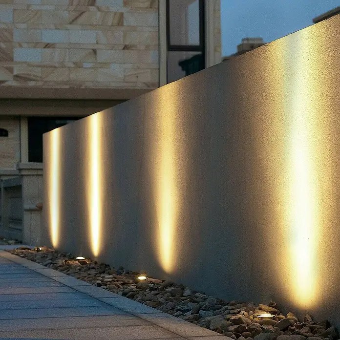 Lucande LED-Gartenstrahler Ava -  - Eingelassene Bodenleuchten werfen warme, vertikale Lichtstrahlen auf eine strukturierte Wand, eingebettet in Kiesel neben einem gepflasterten Weg.