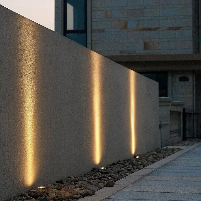 Lucande LED-Gartenstrahler Kenan - 3.000 K - Warme Bodenstrahler werfen vertikale Lichtstrahlen auf eine strukturierte Wand, eingebettet in ein Steinbeet neben einem gepflasterten Weg.