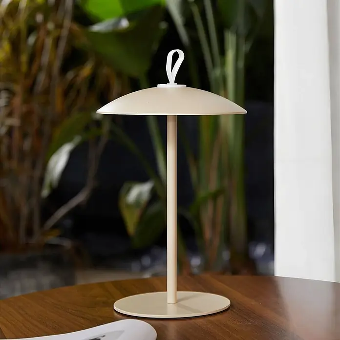 Lindby LED-Tischleuchte Kanani - L x B x H: 18,1 x 18,1 x 30,8 cm, creme / amber, creme / amber, Warmweiß - Beige tragbare Tischlampe mit schirmförmigem Schirm und Trageschlaufe auf einem Holztisch.