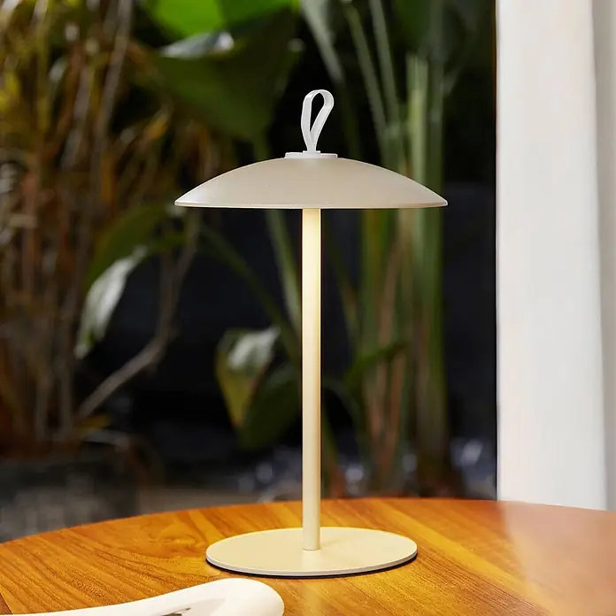 Lindby LED-Tischleuchte Kanani - L x B x H: 18,1 x 18,1 x 30,8 cm, creme / amber, creme / amber, Warmweiß - Eine cremefarbene, moderne Tischlampe mit schirmartigem Schirm und oberer Schlaufe, beleuchtet auf einem Holztisch.