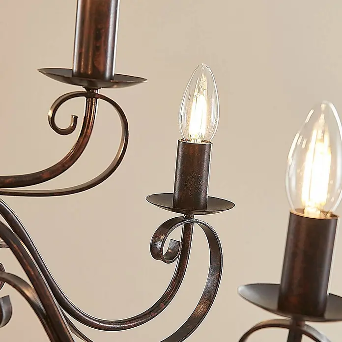 Lindby Kronleuchter Caleb - braun / rost, rostbraun, E14 - Detailansicht eines rustikalen Bronzeleuchters mit Schnörkeln und beleuchteten Kerzen-LED-Lampen.