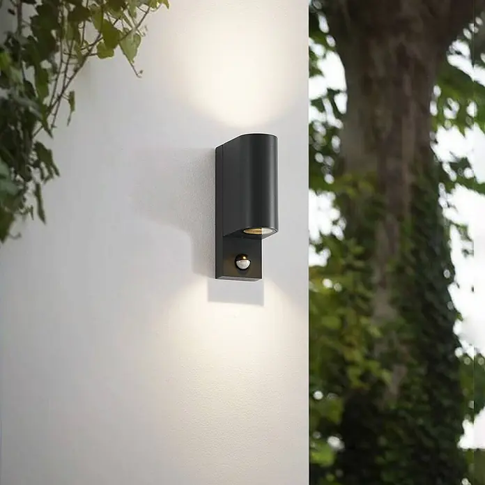 Lindby Außenwandleuchte Palina - 12 x 7 x 21,6 cm, schwarz, IP44 - Schwarze Außenwandleuchte mit Ober-/Unterlicht und integriertem Bewegungsmelder, an weißer Wand zwischen Laub.