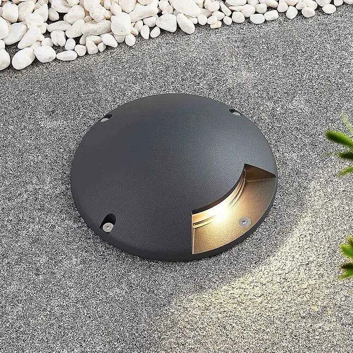 Lindby LED-Gartenstrahler Cormac - 3.000 K - Runde dunkelgraue Bodeneinbauleuchte mit warmem Licht, in einen gesprenkelten Steinweg integriert.