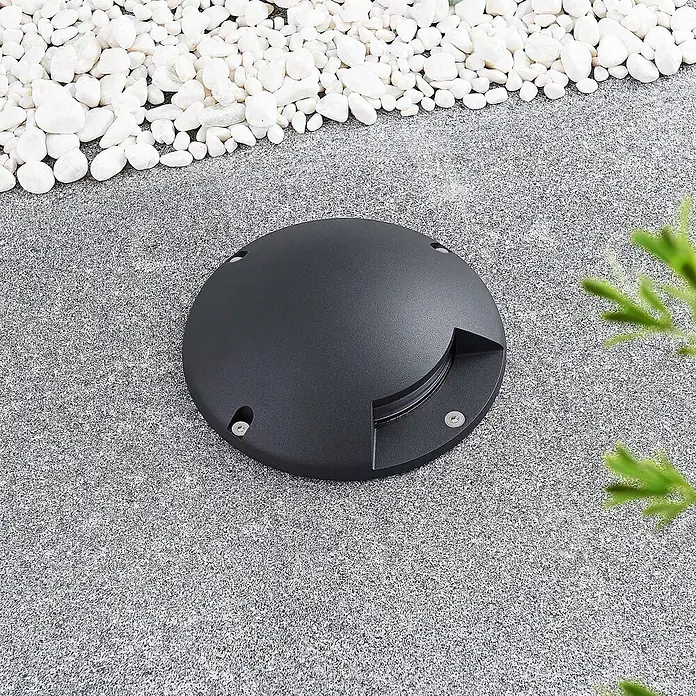 Lindby LED-Gartenstrahler Cormac - 3.000 K - Runde schwarze Bodeneinbauleuchte, in gesprenkelter grauer Oberfläche neben weißen Kieselsteinen.