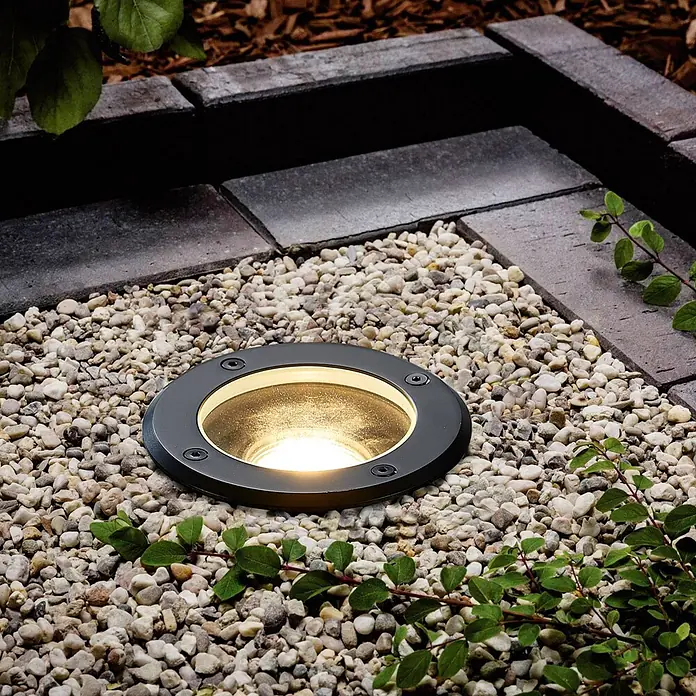 Lindby LED-Gartenstrahler Cliff -  - Schwarze runde Bodeneinbauleuchte beleuchtet helle Kieselsteine auf einem Gartenweg neben dunklen Pflastersteinen.
