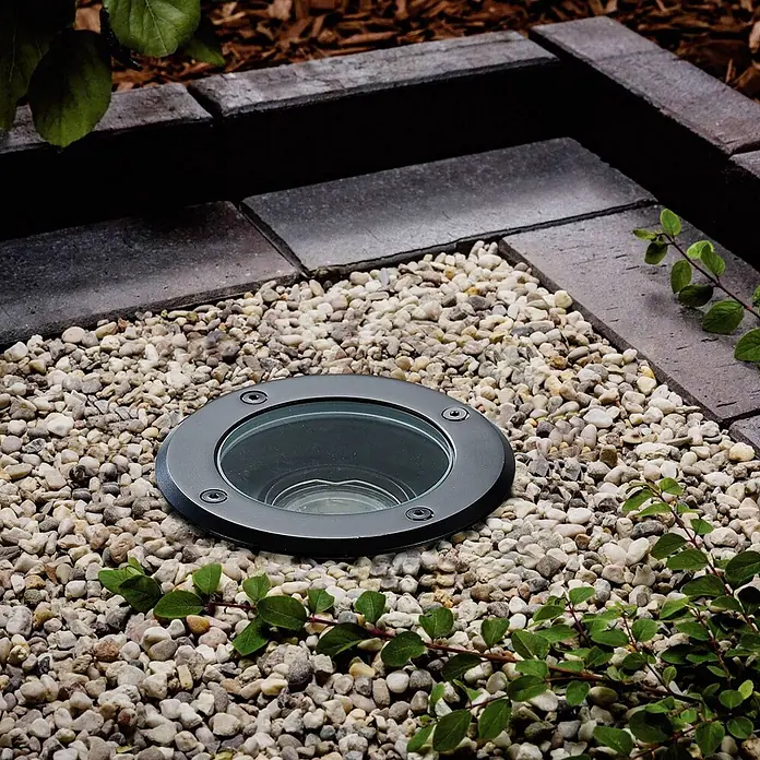 Lindby LED-Gartenstrahler Cliff -  - Schwarze runde Bodeneinbauleuchte mit Glasoberfläche, in hellem Kies neben dunklen Pflastersteinen und Grünzeug.