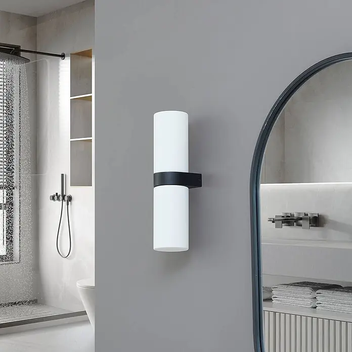 Lucande Wandleuchte Basolo - weiß / opal, E14 - Vertikale zylindrische Wandleuchte aus weißem Glas mit schwarzem Band, die ein modernes Badezimmer beleuchtet.