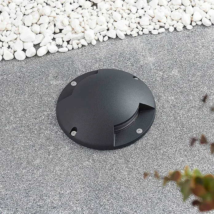 Lindby LED-Gartenstrahler Cormac - 3.000 K - Runde dunkelgraue Bodenleuchte, im Pflaster neben weißen Kieselsteinen eingelassen.