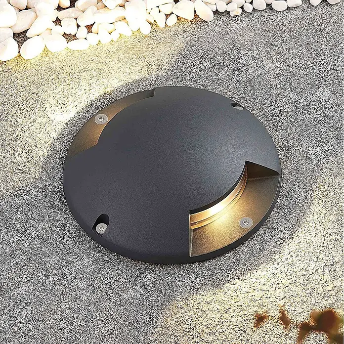 Lindby LED-Gartenstrahler Cormac - 3.000 K - Dunkelgraue runde Bodeneinbauleuchte beleuchtet granular gepflasterten Weg und weiße Kieselsteine warm.