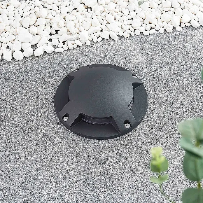 Lindby LED-Gartenstrahler Cormac - 3.000 K - Schwarze runde Bodeneinbauleuchte, bündig in Außenpflaster montiert, neben weißen Steinen.