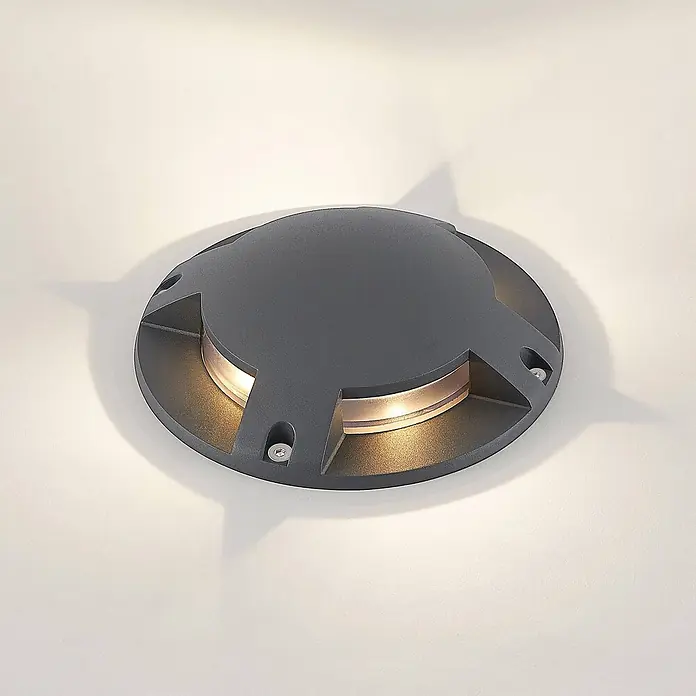 Lindby LED-Gartenstrahler Cormac - 3.000 K - Dunkelgraue runde LED-Bodeneinbauleuchte, die warmes Licht aus seitlichen Öffnungen abstrahlt.