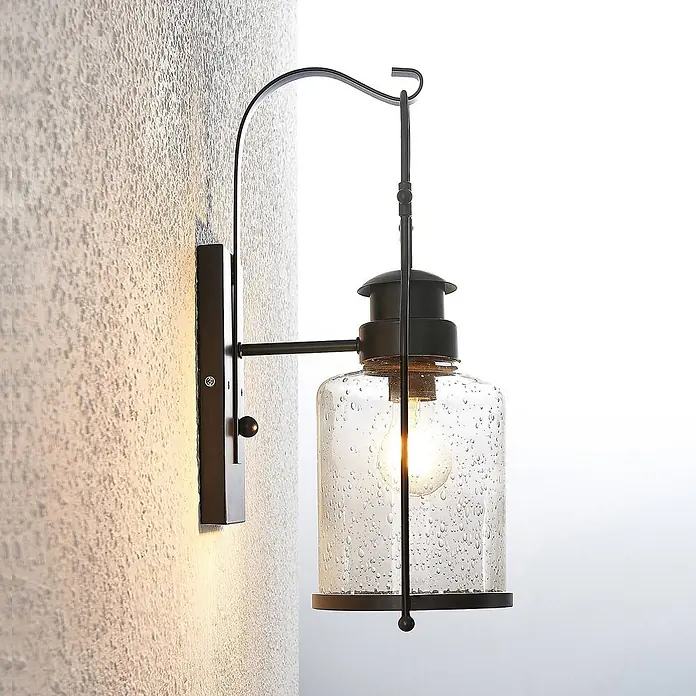 Lindby Wandleuchte Rozalie - L x B x H: 22 x 18 x 44 cm, schwarz, E27 - Schwarze Außenwandleuchte aus Metall mit geschwungenem Arm und zylindrischem, blasigem Glas, das eine leuchtende Glühbirne umschließt, an einer strukturierten Wand montiert.