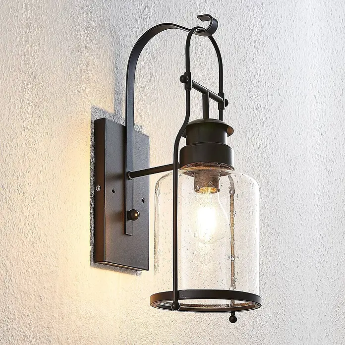Lindby Wandleuchte Rozalie - L x B x H: 22 x 18 x 44 cm, schwarz, E27 - Dunkle Metall-Wandleuchte mit zylindrischem Schirm aus strukturiertem Klarglas und sichtbarer Glühbirne, an strukturierter Wand montiert.