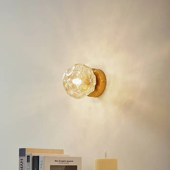 Lindby Wandleuchte Piatra - L x B x H: 12,5 x 15,5 x 15,5 cm, creme / amber, G9 - Wandleuchte mit strukturierter Glasschirm und Messingfuß, beleuchtet eine helle Wand über Büchern und einer Kerze.