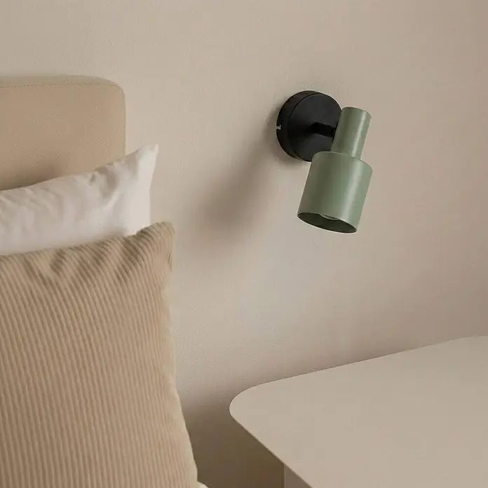 Lindby Wandstrahler Ovelia - schwarz, grün, E27 - Eine matte grüne Wandleuchte mit schwarzem Sockel an einer Wand neben einem Bett mit Kissen und einem hellen Nachttisch.
