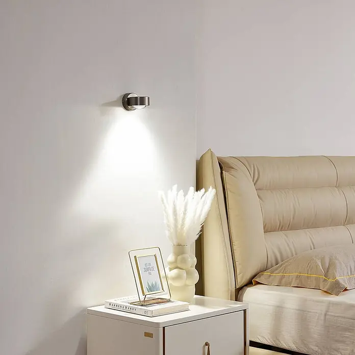 Lindby Wandleuchte Jyla - L x B x H: 10,5 x 8 x 7 cm, alu / grau / zink, 2GX53 - Dunkelgraue Wandleuchte über einem Nachttisch, die ein warmes Licht nach unten wirft.
